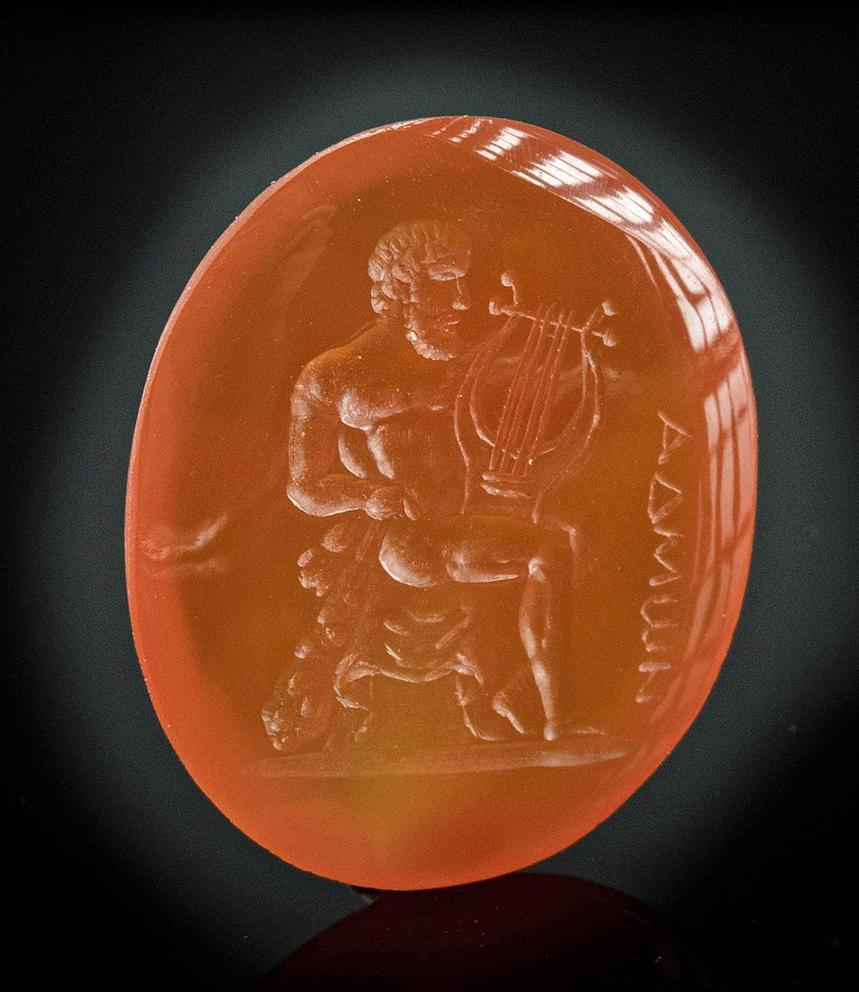 Neoclassical Carnelian Intaglio, Nude Male & Greek Text: Western Europe, Neoclassical Period, ca. late 18th to 19th century CE. A finely preserved carnelian intaglio of an ovoid form depicting a nude male seated upon a stool. The man bends his left leg slig