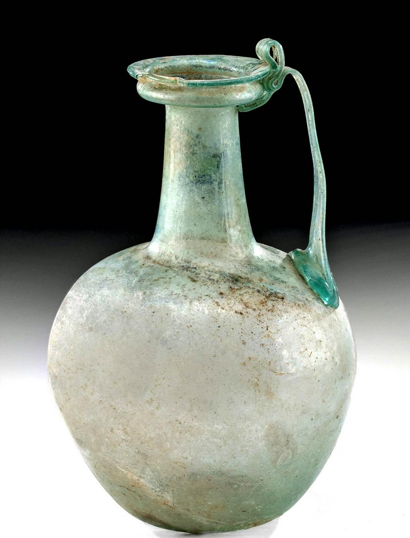 Roman Glass Pitcher w/ Elegant Trailed Handle: Roman, Imperial Period, ca. 1st to 3rd century. CE. This beautiful free-blown glass pitcher sits upon a concave base and rises into a globular body with a long cylindrical neck and collared rim. A tra