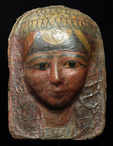 An Egyptian Cartonnage Painted Funerary Mask: New Kingdom, ca. 1500 BC. Linen and plaster / gesso mask, finely-decorated; wig painted with a lotus blossom and flesh tone face with almond shaped eyes in white and black pigment outlines the pupils