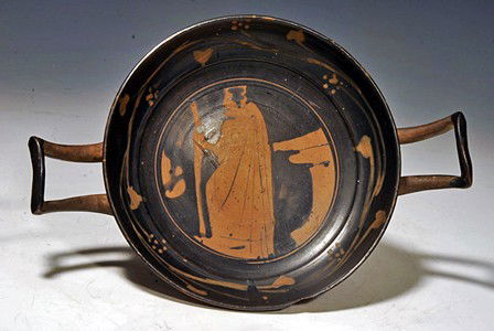 A Greek Attic Stemless Kylix Attributed to YZ Group: Athens, ca. 370 to 350 BC. The tondo decorated with a man wearing the himation and holding a staff in his hand, standing next to an altar, framed by a wreath of ivy and grapes. The exterior depicting