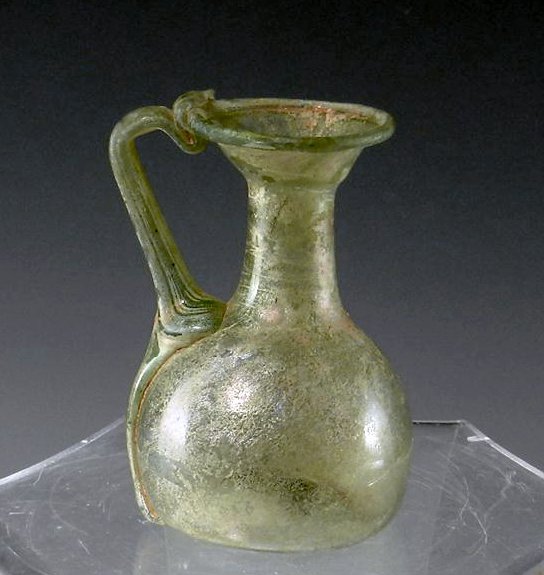 A Roman Glass Handled Juglet: Rome, Ca. 2nd Century CE. Handle spans the whole body, attaching at the rim and attaching at the top of body, like a traditional handle, but then the glass handle bleeds down to the base. Beaker takes