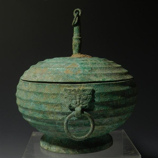 An Archaic Chinese Zhou Dynasty Ritual Food Vessel: China, Early Western Zhou Dynasty, 10th Century BC . Bronze vessel stands on a raised platform; body ribbed by concentric horizontal circle pattern. Single handle is in form of applied Taotie mask wit