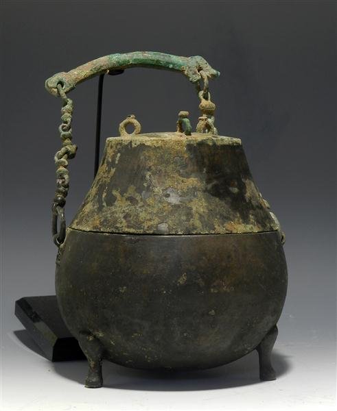 A Han Dynasty Bronze Hou-Lou Vessel: China, Western Han period (206 BC - AD 9). A cast-bronze carinated cauldron base with tripod feet and removable top, two staples with suspension chain formed with double-loop links and broad central h