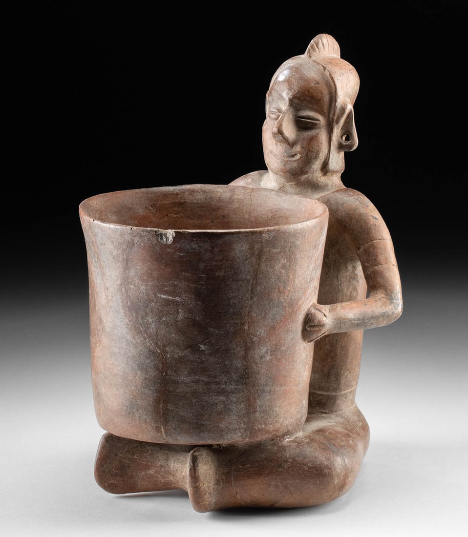 Colima Redware Vessel - Seated, Smiling Figure: **Originally Listed At $600** Pre-Columbian, West Mexico, Colima, ca. 300 BCE to 300 CE. A charming redware vessel in the form of a seated, smiling figure wearing a crested helmet as he holds a giant,