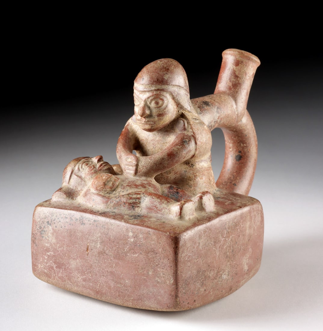 Moche Pottery Vessel w/ Medical or Ceremonial Scene: Pre-Columbian, North Coast Peru, early Moche, Period I, ca. 100 BCE to 300 CE. A captivating Moche redware stirrup vessel depicting a compelling scene: two figures engaged in what appears to be a medi