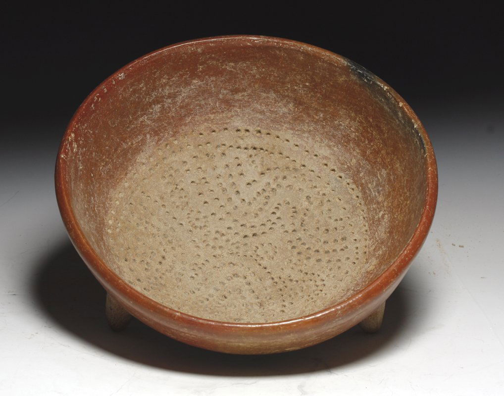 A Chupicuaro Tripod Grater in Center, ex-Wray: Pre-Columbian, West Mexico, Chupicuaro culture, ca. 300 BCE. Red slipped pottery tripod vessel with some black paint marks on exterior. Inside is incised with circular indents in a "cheese grater" pat