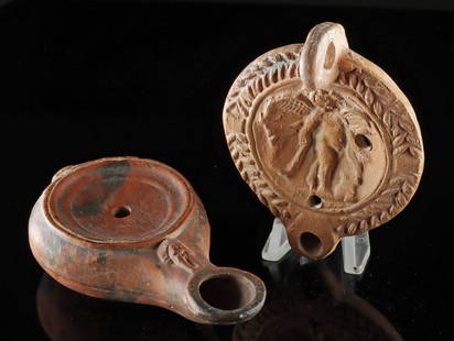 2 Roman Oil Lamps - Cupid w/ Laurel Tondo + Slender Form: Roman, Imperial period, ca. 1st to 3rd century CE. A delightful duo of pottery oil lamps, each a distinct expression of ancient craftsmanship. The first, sculpted from a deep red clay, presents a slen