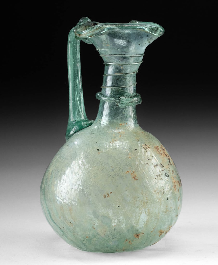 Elegant Ancient Roman Trailed Glass Pitcher: Roman Empire, ca. 1st to 3rd century CE. A beautiful pale green blown glass pitcher with a globular body featuring faint vertical ribbing, a cylindrical neck with applied trailing, and a wide flared m