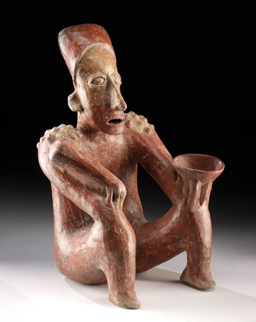 Jalisco Seated Nude Male Figure with Bowl and Mace: Pre-Columbian, West Mexico, Jalisco, ca. 300 BCE to 300 CE. A powerfully modeled seated male figure from ancient Jalisco, sculpted in buff clay with a rich red slip and burnished finish. Rendered in t