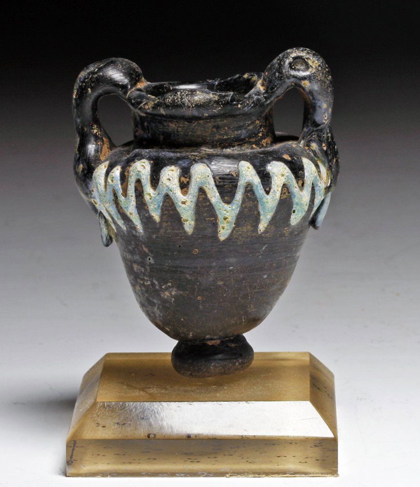 Islamic Coreform Glass Amphora: Middle East, Ca. 7-9th Century CE. Miniature ancient vessel in dark blue/black glass sits on raised glass base; body and handles form traditional Greek amphora shape. Painted blue glass zigzag pattern