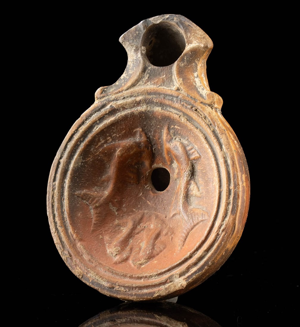 Roman Molded Oil Lamp - Twin Dolphins in Relief: Roman, Imperial Period, ca. 1st to 3rd century CE. A terracotta oil lamp, mold-formed featuring a recessed discus decorated with two stylized dolphins in relief. The spout projects outward with a roun