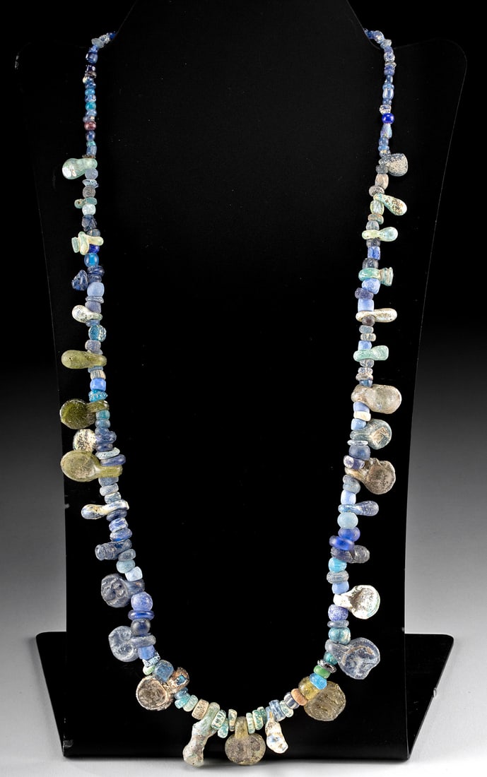 Roman-Byzantine Glass Bead Necklace & Stamped Pendants: Roman, Imperial Period to Byzantine Periods, ca. 1st to 7th century CE. A luminous necklace composed of ancient glass beads in shades of cobalt, aqua, and green, strung on modern monofilament wire. Al