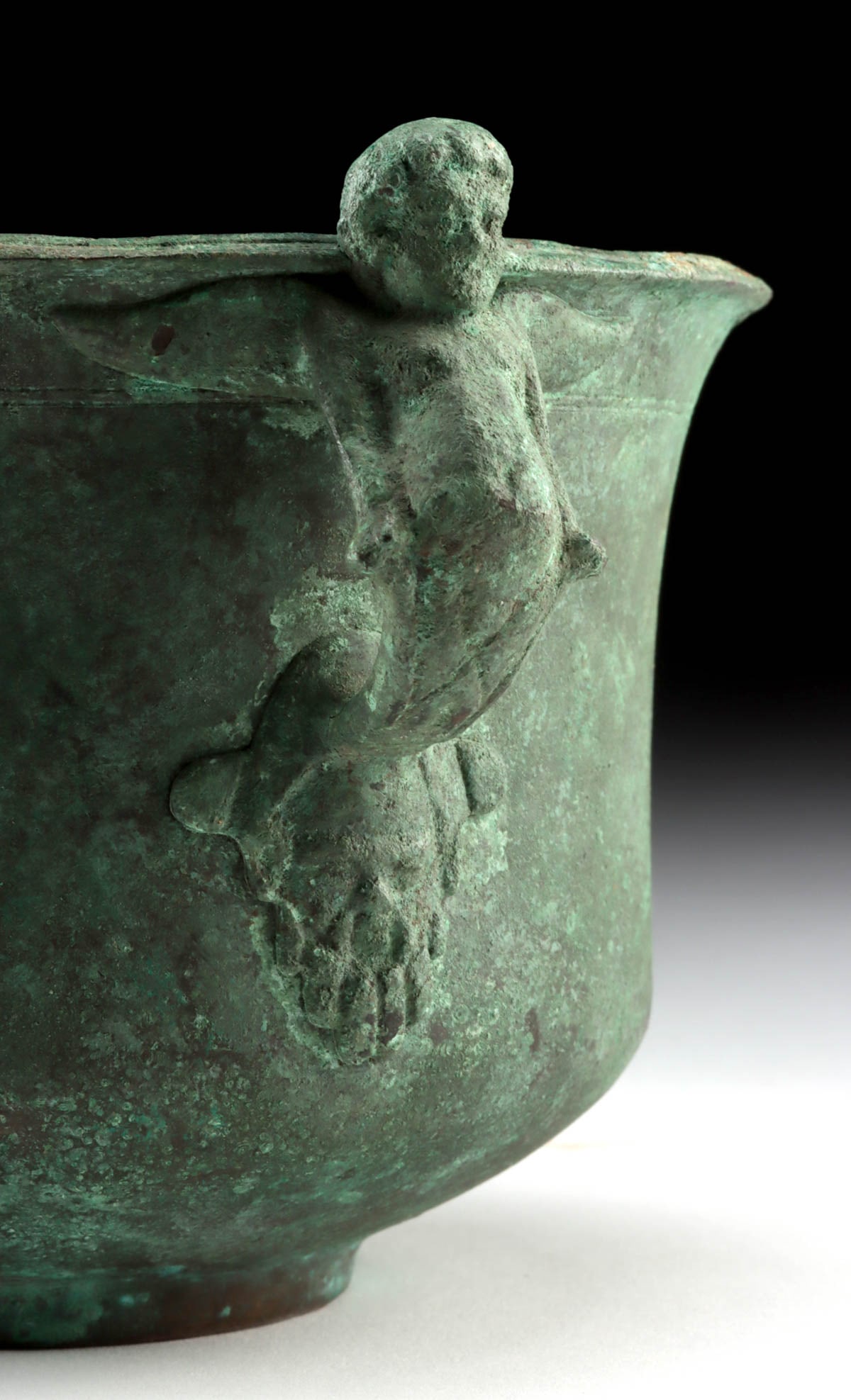 Roman Bronze Drinking Vessel w/ Winged Eros Handle: Roman, Imperial Period, ca. 1st to 3rd century CE. A bronze vessel with a rounded body rising from a low foot and opening to a gently flared rim, now coated in a deep green patina. At the side is an a