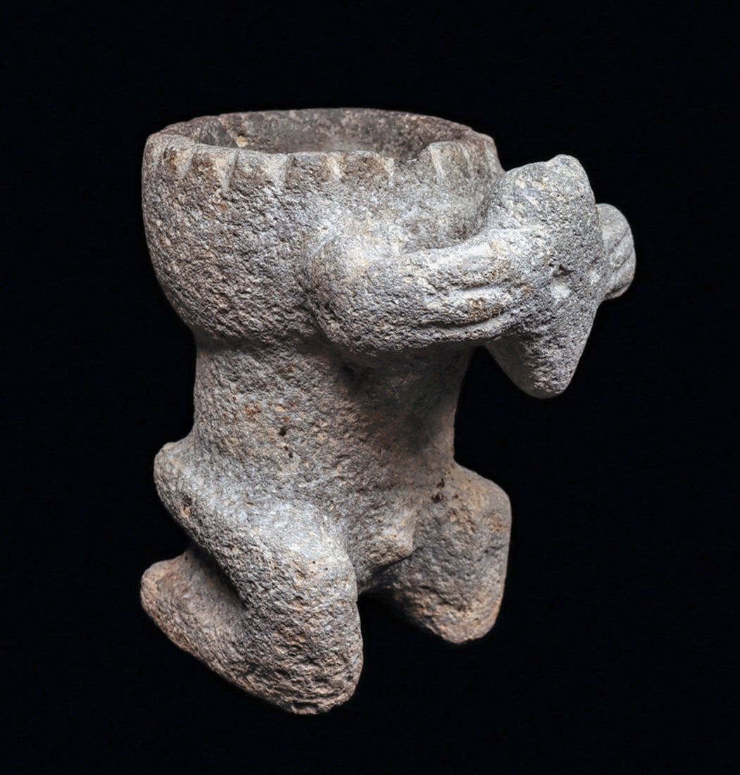 Jalisco Kneeling Transformational Figure with Bowl: Carved gray basalt kneeling male shaman figure with a large bowl on his shoulders, having a segmented rim decoration, and holding both hands to his animal-form head. Overall scattered areas of surface