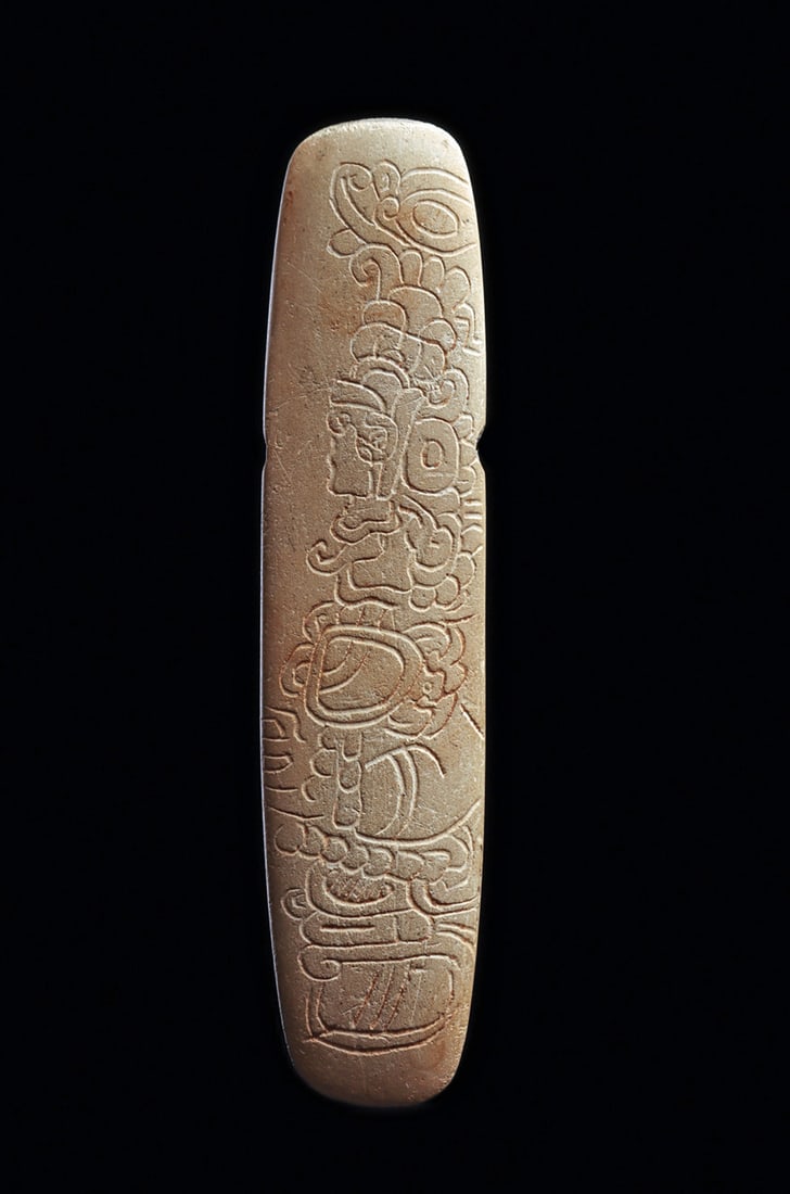 Maya Limestone Decorated Celt Pendant: Carved compacted limestone celt form pendant, having a biconical drilled suspension hole. Incised elaborate decoration on the front depicting a chief seated on top of a deity. Good ancient tan-brown s