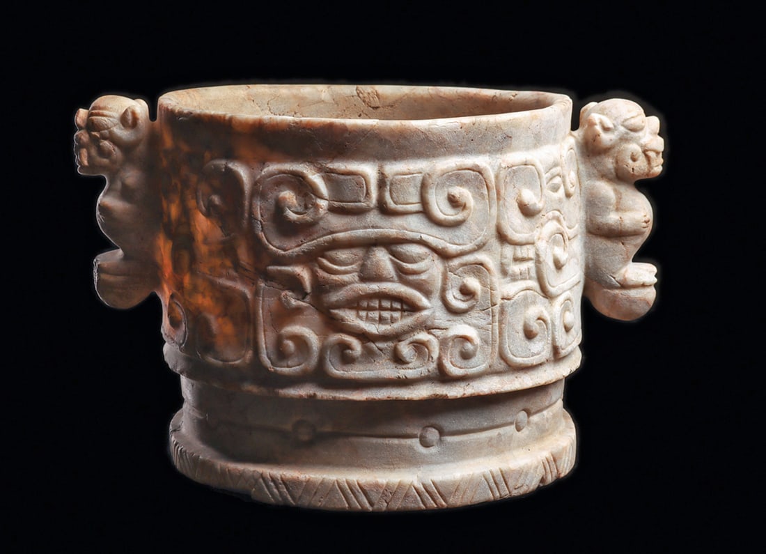 Ulua Valley Maya Marble Vase: Carved semi-translucent limestone-marble cylindrical vessel carved on each side with a stylized zoomorphic head with lunate eyes, ovoid mouth showing teeth, with opposed volutes on the forehead. Surro
