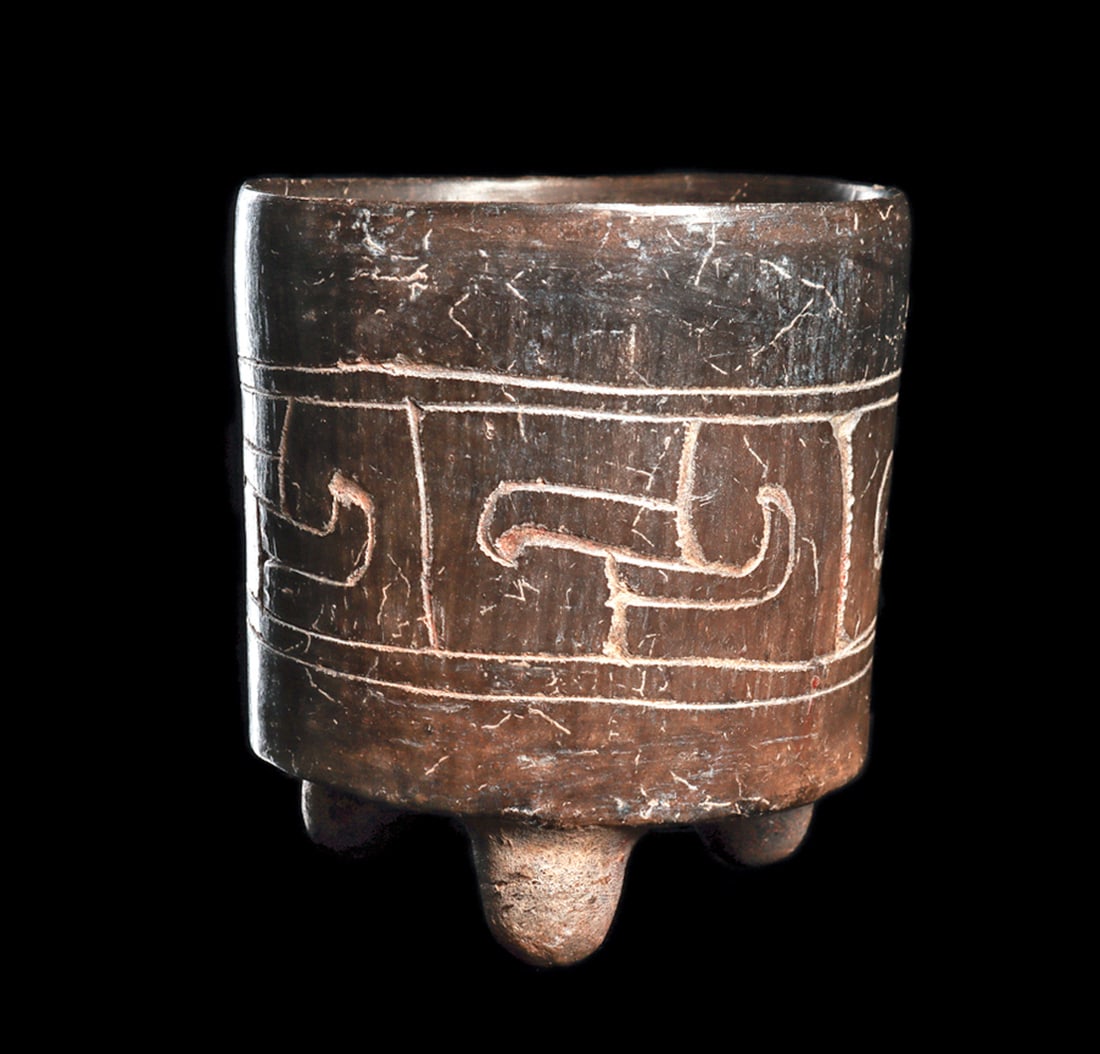 Maya Blackware Tripod Vessel with Glyph: Blackware pottery small vase with short tripod legs, decorated with an incised wide center band depicting four abstract profile bird heads. Scattered areas of root marks and surface deposits. Two mino