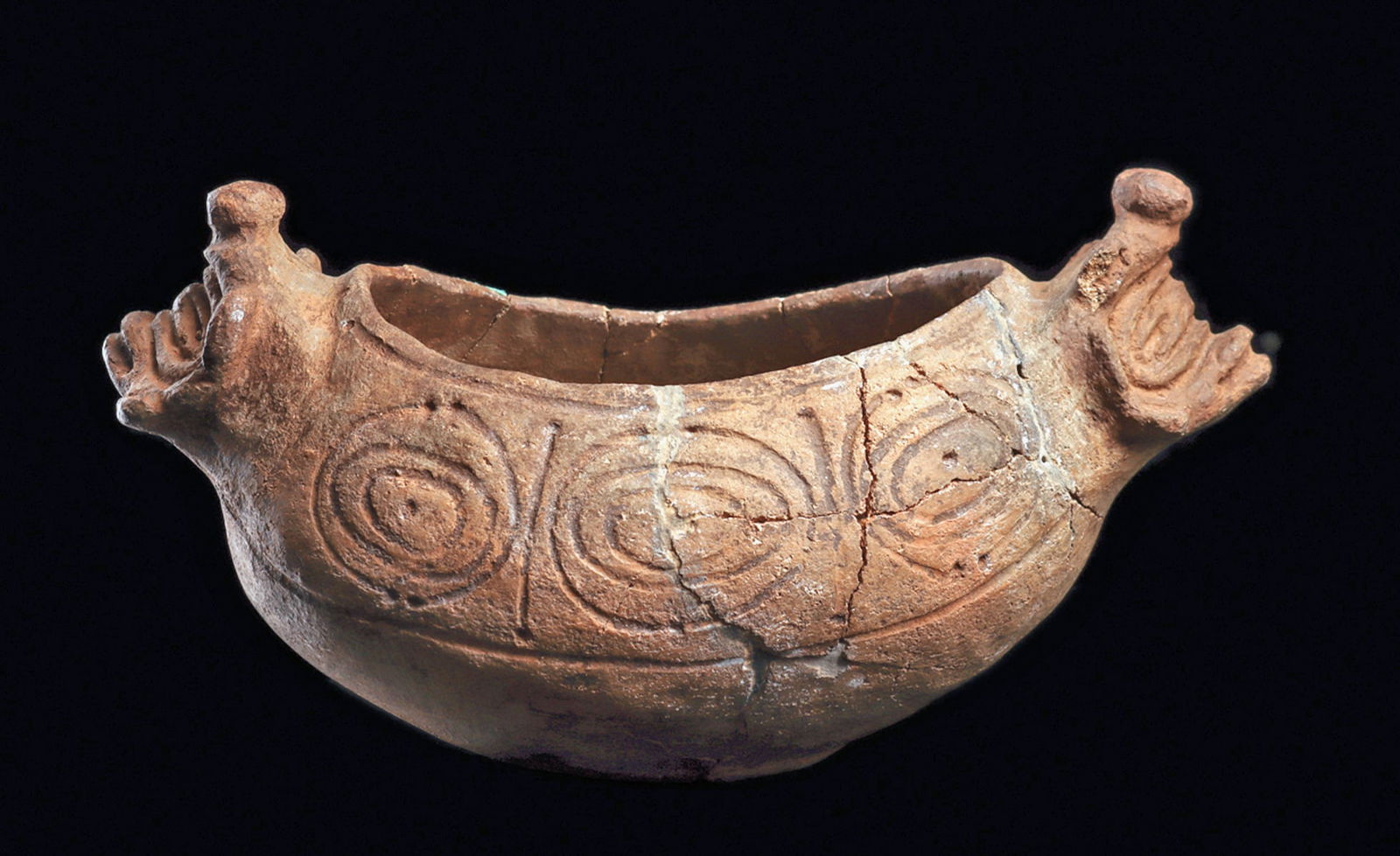 Taino Decorated Vessel: Brown ware pottery oblong bowl having opposing stylized deity heads at both ends. Decorated with a wide upper body bands with three concentric ring elements on both sides. Repaired from approximately