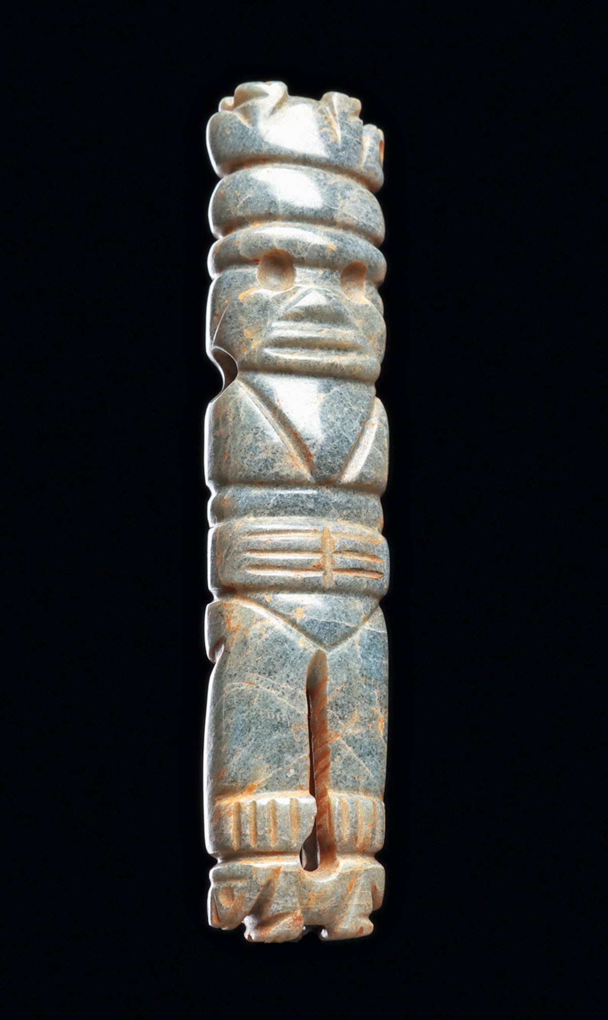 Costa Rican Jade Figural Pendant: Carved blue-gray jade anthropomorphic pendant depicting a standing figure holding both hands over his stomach, with delineated fingers. Wearing a tall headdress with an animal lying on it back at the