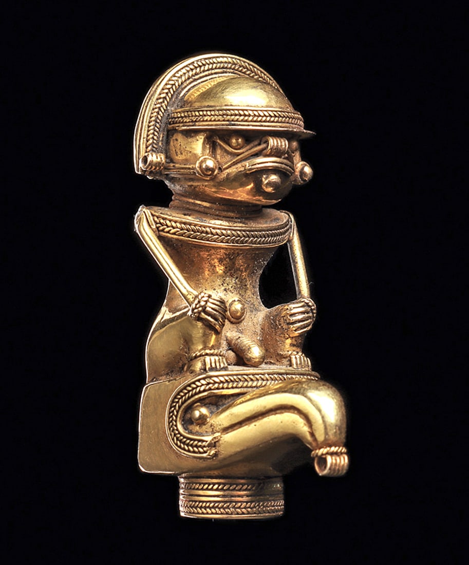 Tairona Gold Cacique Pendant: Fine gold male cacique pendant, seated on top of a bird's head, with both hands resting on his knees. Wearing a fancy dome helmet with two faux braided borders, two small integral suspension loops on