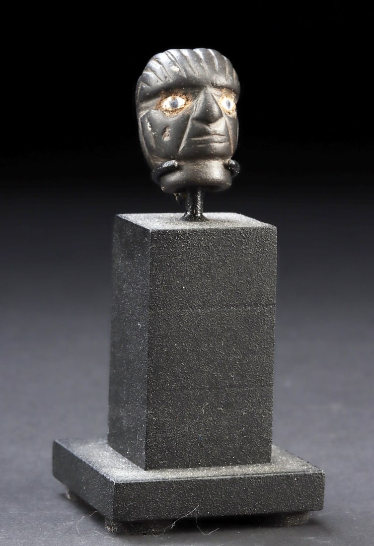 Miniature Moche Stone Face w/ Inlaid Shell Eyes: Pre-Columbian, North Coast Peru, Moche, ca. 2nd to 7th century CE. A miniature head carved from a black stone with a dramatic visage comprised of wide-open, iridescent white shell inlaid eyes with bla