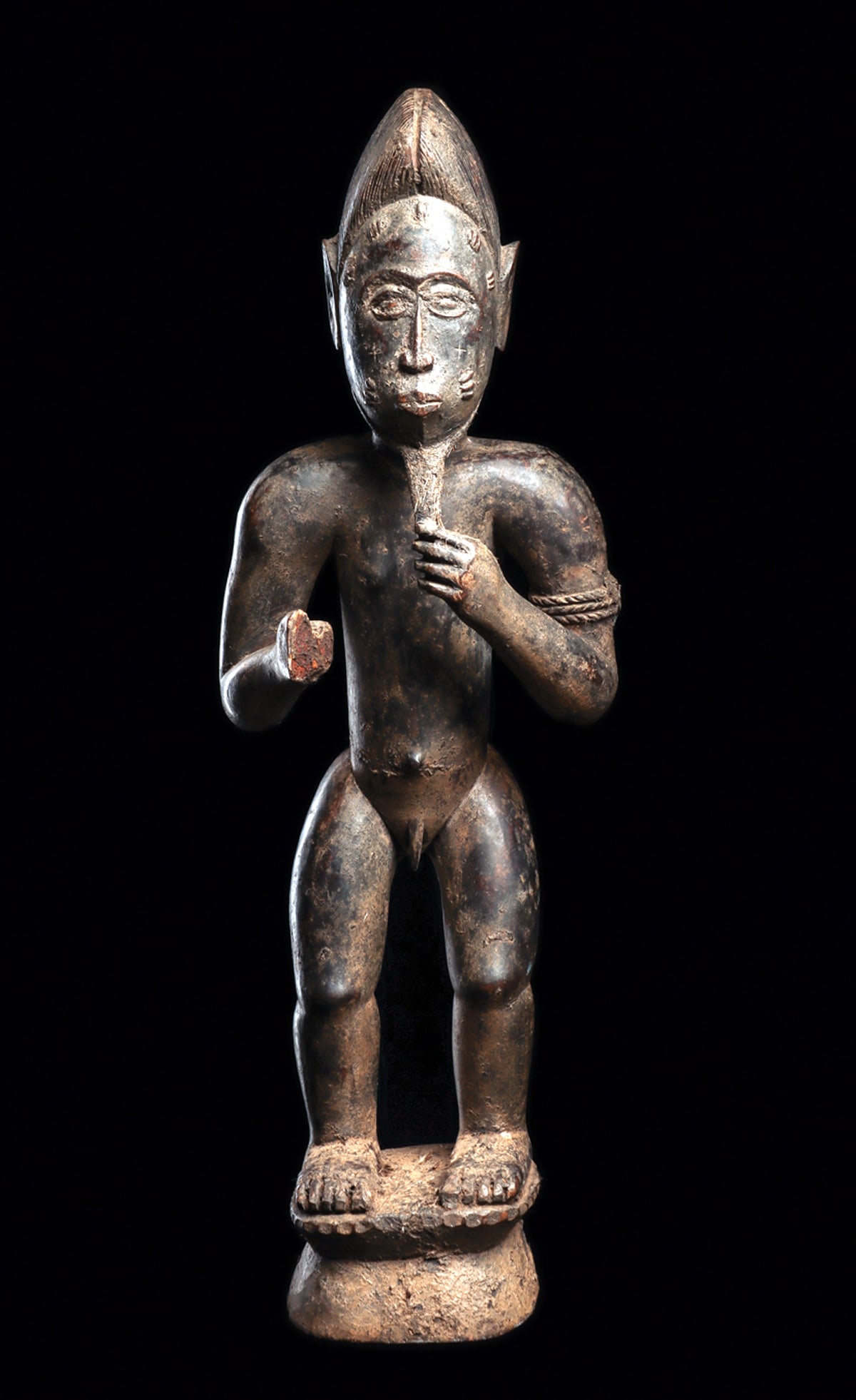 Baule Male Figure: Well carved Baule male figure stands on an integral base. The figure raises his left hand to a long, carefully rendered beard and wears a tall crested coiffure with a short plait at the nape. Large,
