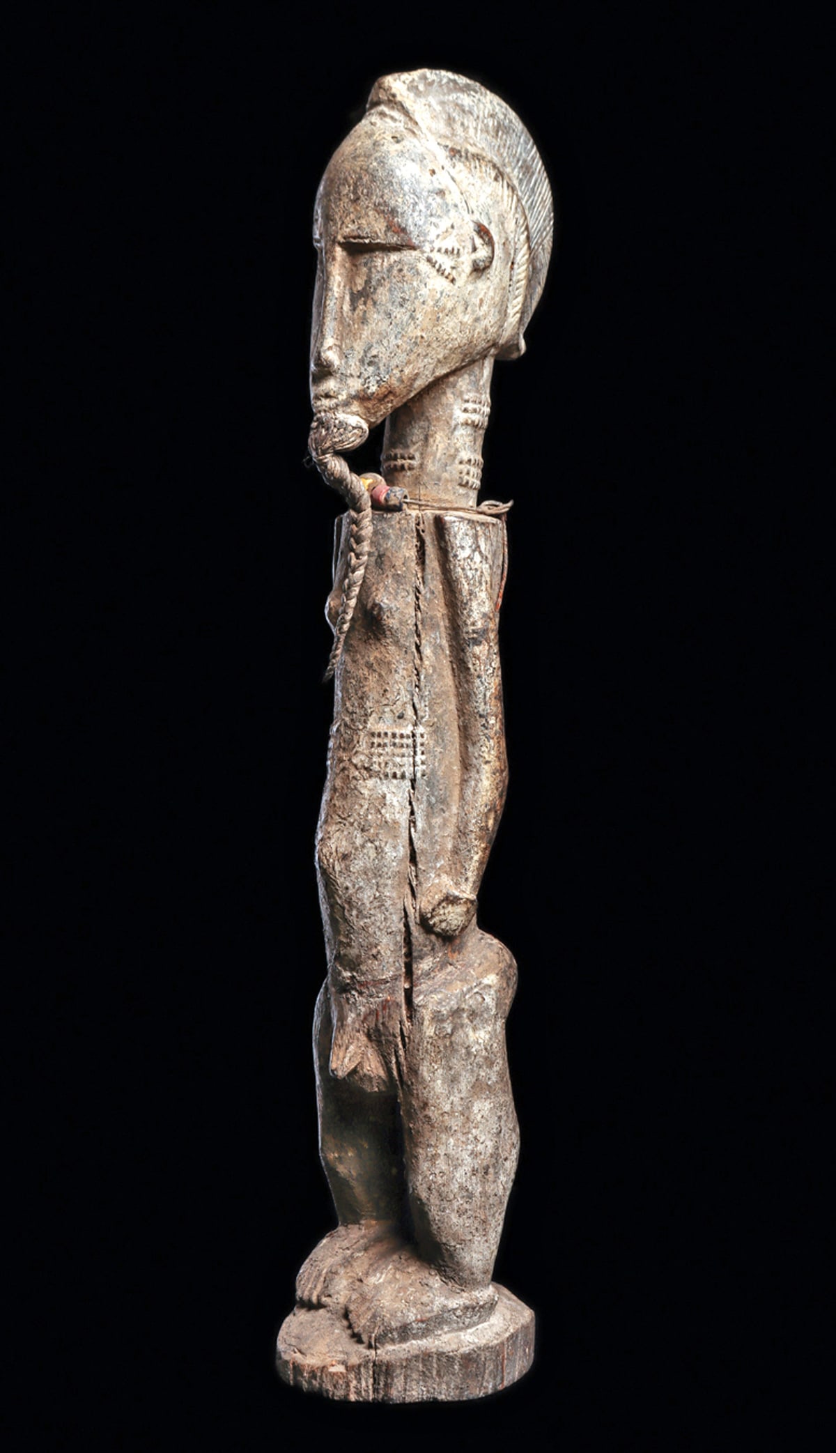 Baule Male Figure: Tall carved male spouse figure with attached plaited fiber beard, classic tapered crest coiffure, scarification around face, neck, torso, long slender relief arms, and adorned with a fiber cord neckla