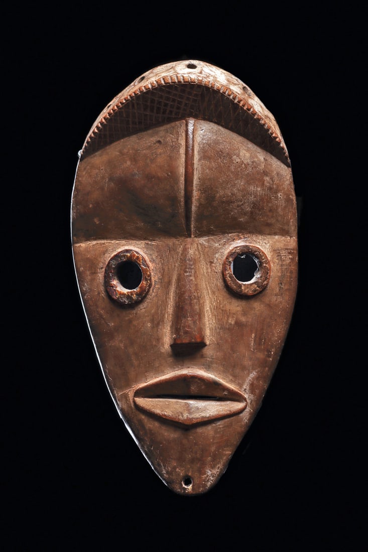 Dan Maskette: Of shallow convex form with short coiffure on top, wide forehead with relief ridge, round rim eyeholes, and wide lips with slit opening. Chin is pierced for attachment. Medium brown burnished patina