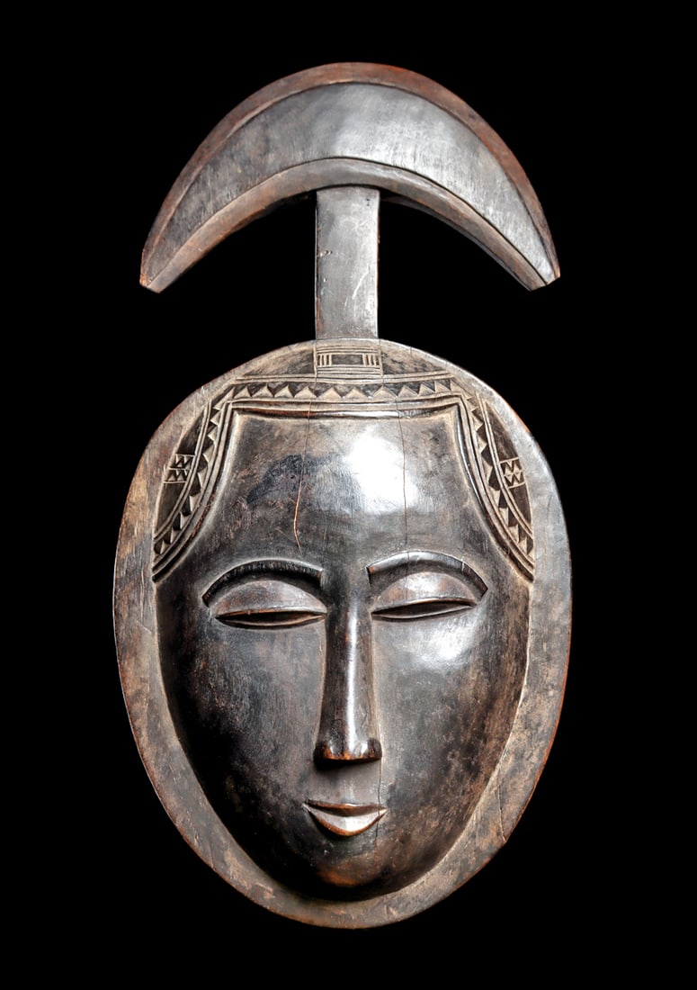 Guro Mask: Carved of hardwood, wide ovoid mask with idealized features, hairdo with geometric elements, and a crescent moon finial rising above. Dark brown smooth patina, with ilght deposits. Hairline age cracks