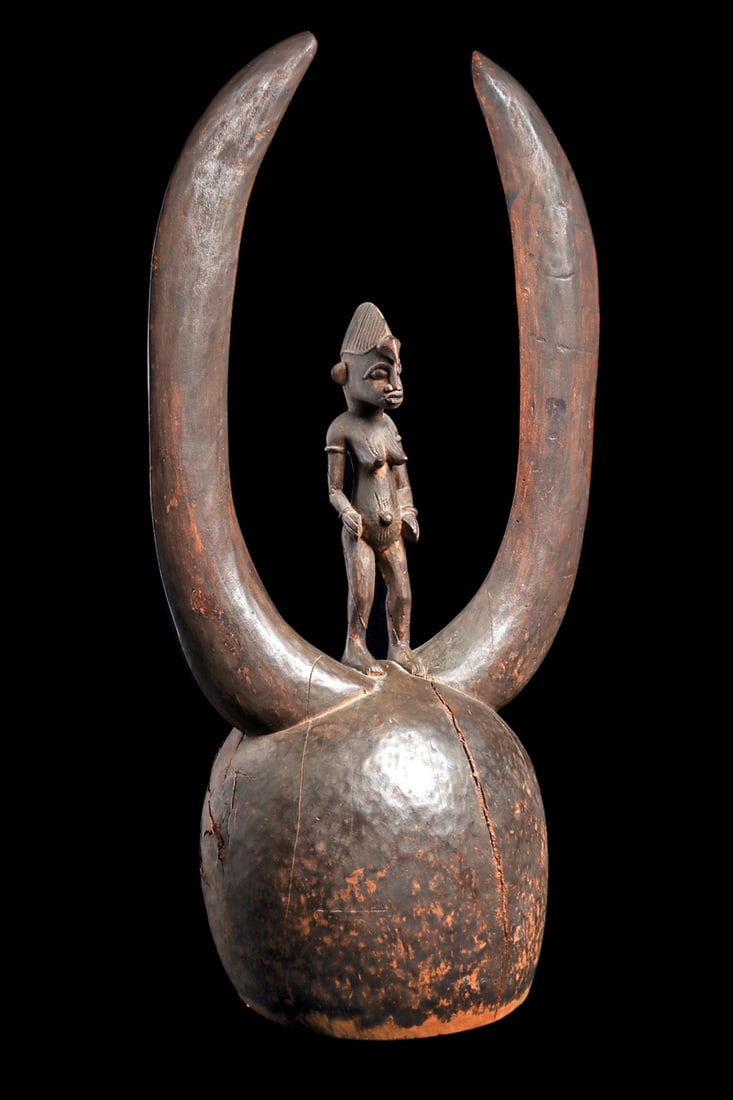 Senufo Helmet Mask: Simple bell like form topped with long plain buffalo horns, standing between them a typical Senufo female figure, cracks in the wood at the back of the mask. 31"H. Provenance: Private collection. Pur