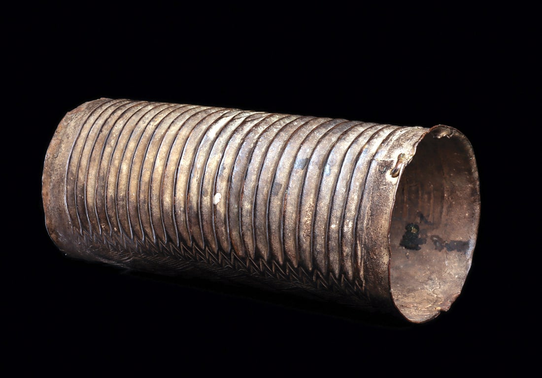 Ibo Bronze Ankle Cuff: Copper alloy tubular leg ornament with continuous ribbed pattern, and engraved vine pattern against crosshatched background along sides of slit opening. Two old native repairs with metal ingots along