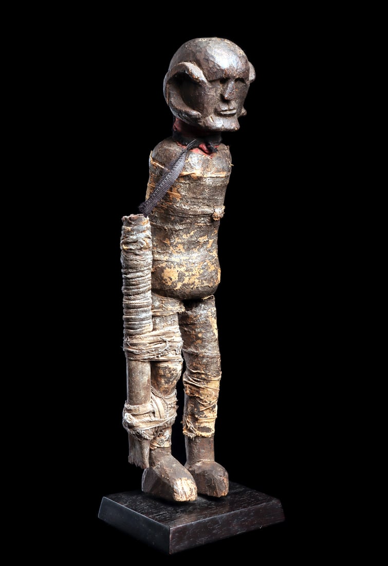 Shambaa Fetish Figure: Fetish figure enveloped in red cloth, now darkened and blackened through age and accumulated ritual deposits. An iron tube is secured to the right leg, reinforcing the figure's charged, empowered char
