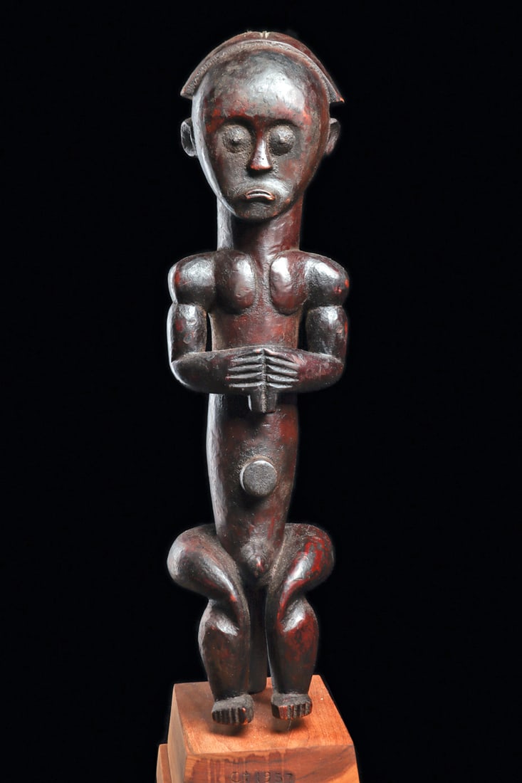 Fang Style Reliquary Figure: Large, decorative, carved wood male figure in style of a reliquary guardian. The elongated figure with small metal nail eyes, holds an object in front of his chest, and sits with rounded hips supporte