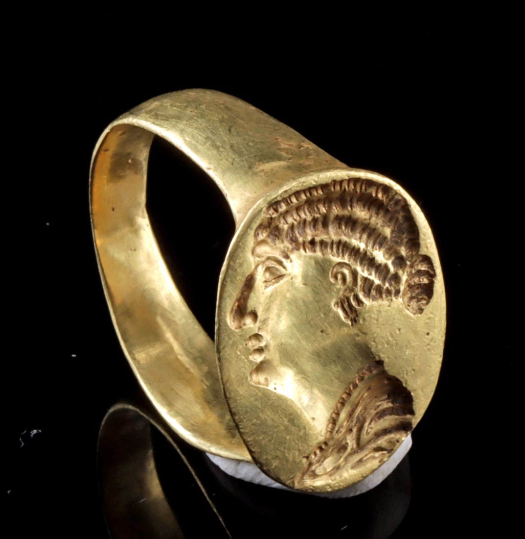 Roman Revival 20 kt Gold Ring w/ Female Portrait: Europe, late 19th–early 20th century CE. The broad hoop surmounted by an oval bezel stamped with a profile portrait bust of a female in the antique manner, the surface with a softly matte finish thr