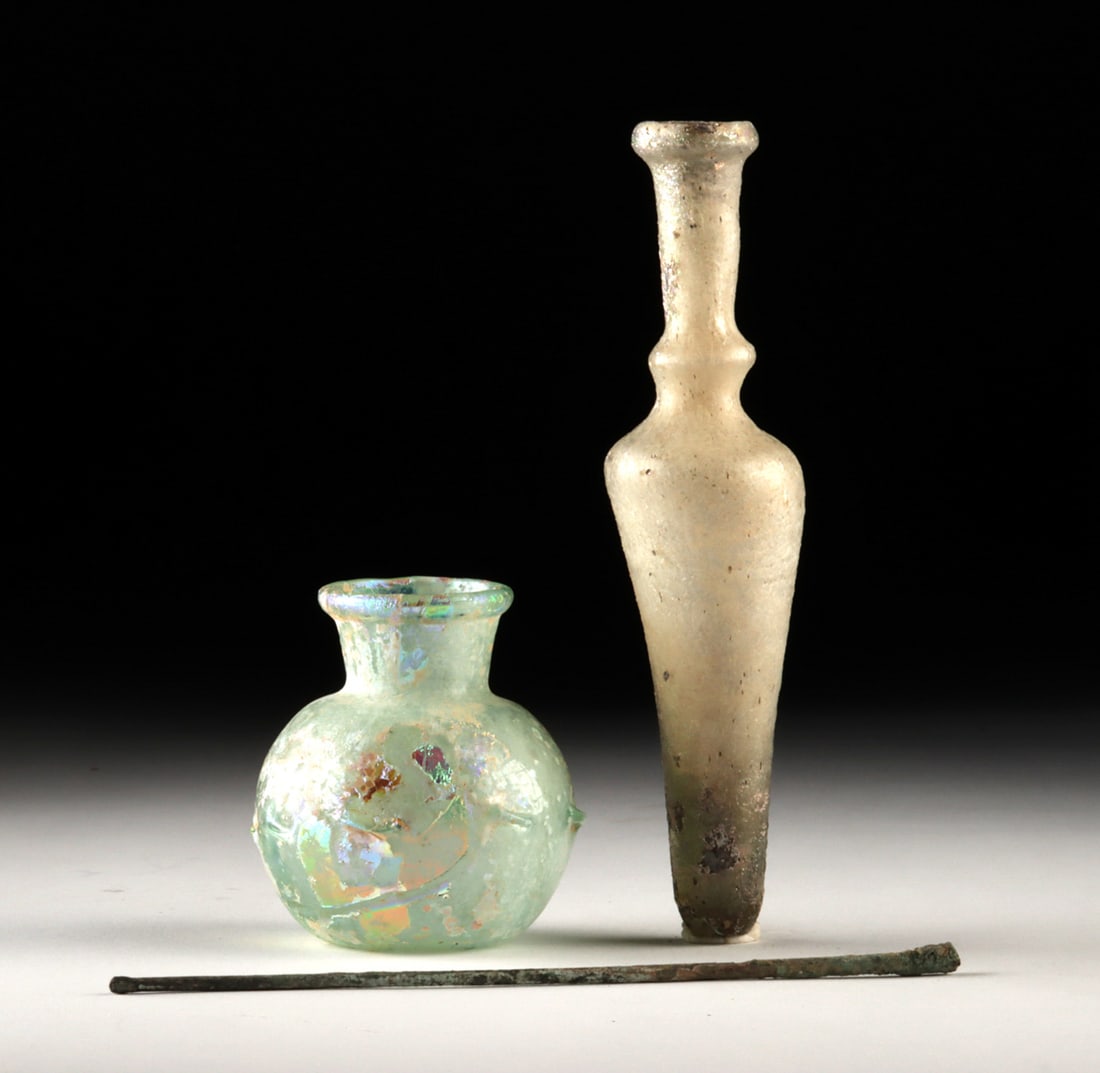 Two Roman Glass Jars + Lead Copper Alloy Applicator: Roman, Imperial period, ca. 1st to 3rd century CE. An alchemy of glass and metal unfolds in this intimate Roman cosmetic group, where translucence meets utilitarian heft and daily ritual leaves its