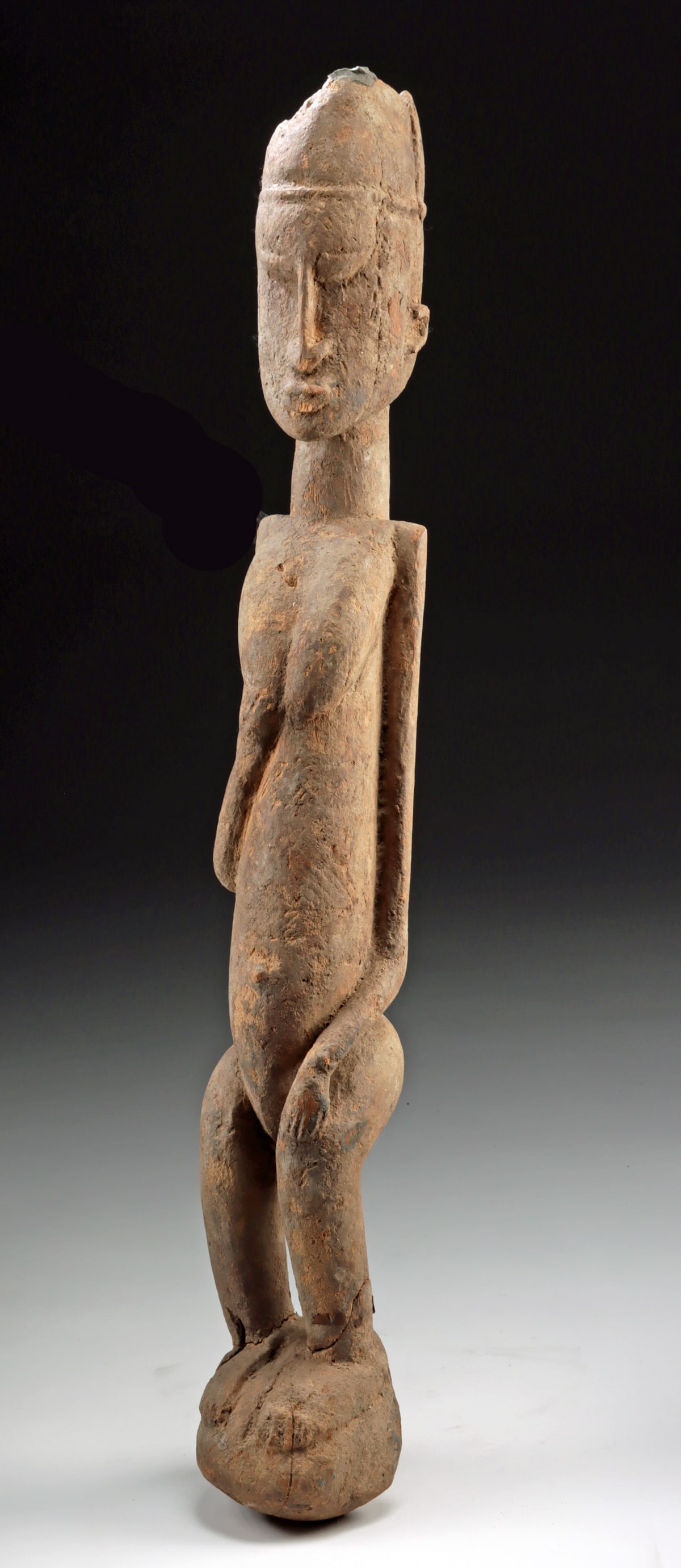 Early 20th C. Mali Dogon Wood Nude Female Figure: West Africa, Mali, Dogon culture, ca. early 20th century CE. A carved Dogon wood figure depicting a standing nude female with legs slightly bent and arms arranged in a modest, inward gesture. One hand
