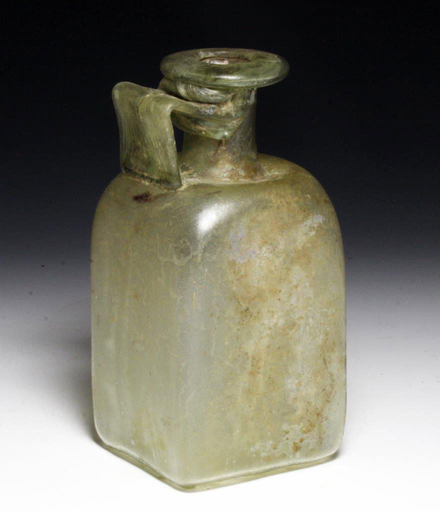 Lg Roman Green Glass Flask with Handle: Rome, ca. 1st - 4th Century CE. A Large Roman glass jug with rectangular body, cylindrical neck, flared rim, and wide textured handle. Sides of body cave slightly in towards center, rim is folded glas
