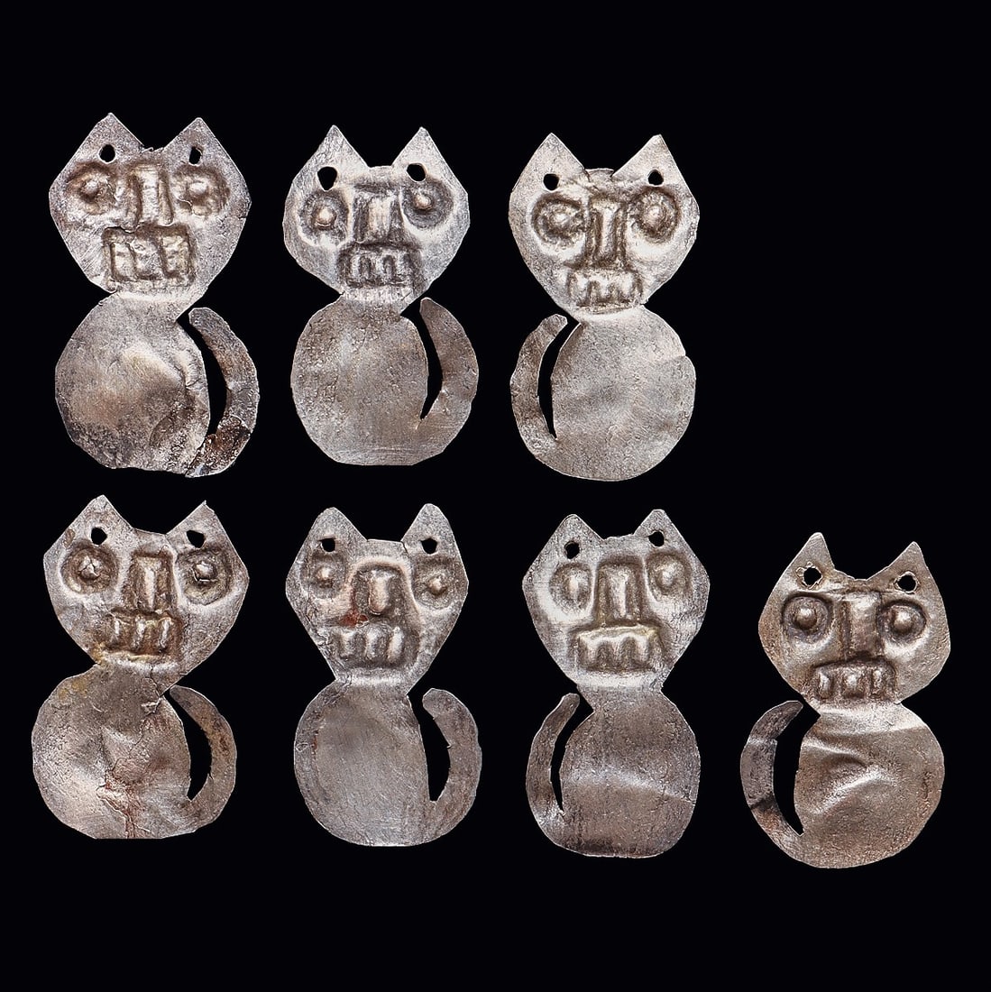 Chimu Silver Feline Dangler Attachments (7): Chimu. Ca. 1000 - 1250 CE. Seven similar sheet silver, shirt danglers, all depicting a feline with a long tail, toothy mouth and perked ears, and having two attachment holes at the top. All have an