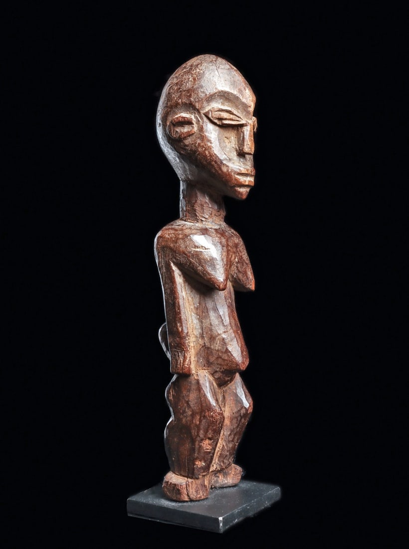 Lobi Female Figure: Burkina Faso. Carved of hardwood, well modeled ovoid head with rimmed eyes, subtle crest, her body with hanging breasts. Losses to feet. Dark brown burnished patina. Custom base. Size: 7.75" H (20cm)