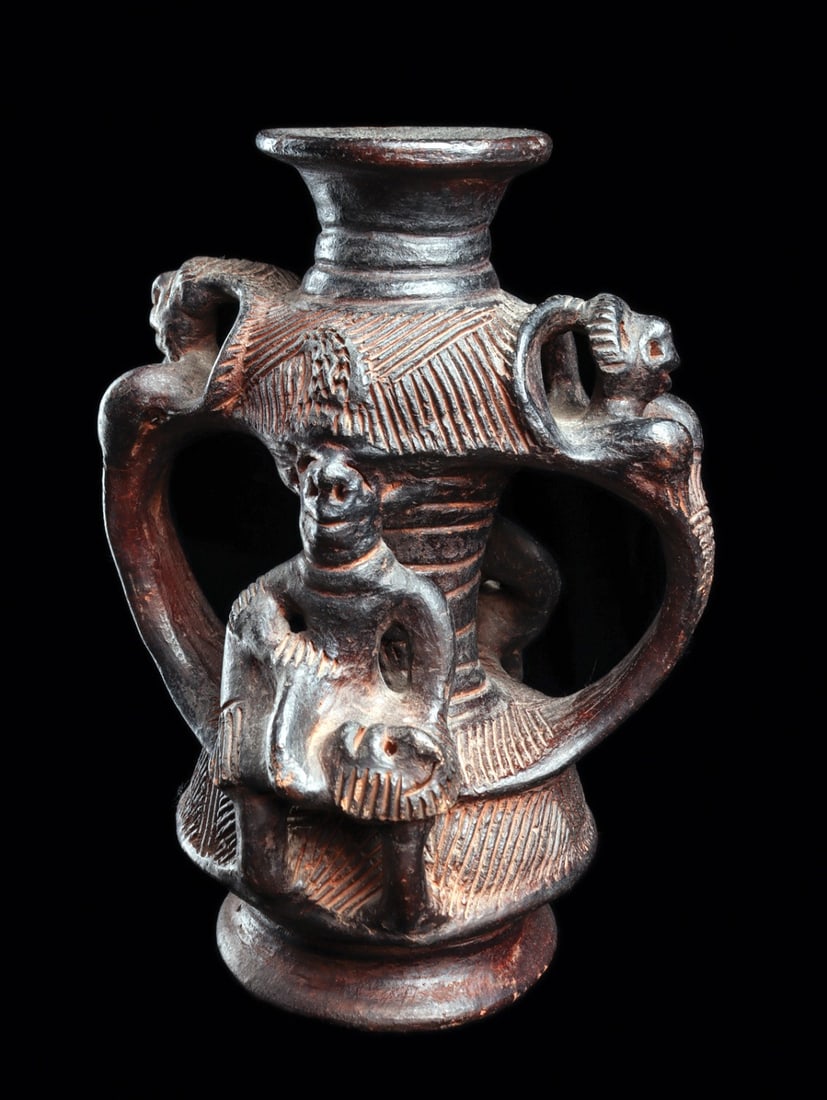 Igbo Maternity Pottery Vessel: Ishiagu, Nigeria. Late 20th century. Blackened glazed terracotta vessel modeled with two figures cradling a child on opposite sides, with two large strap handles each having figural bust with curved a