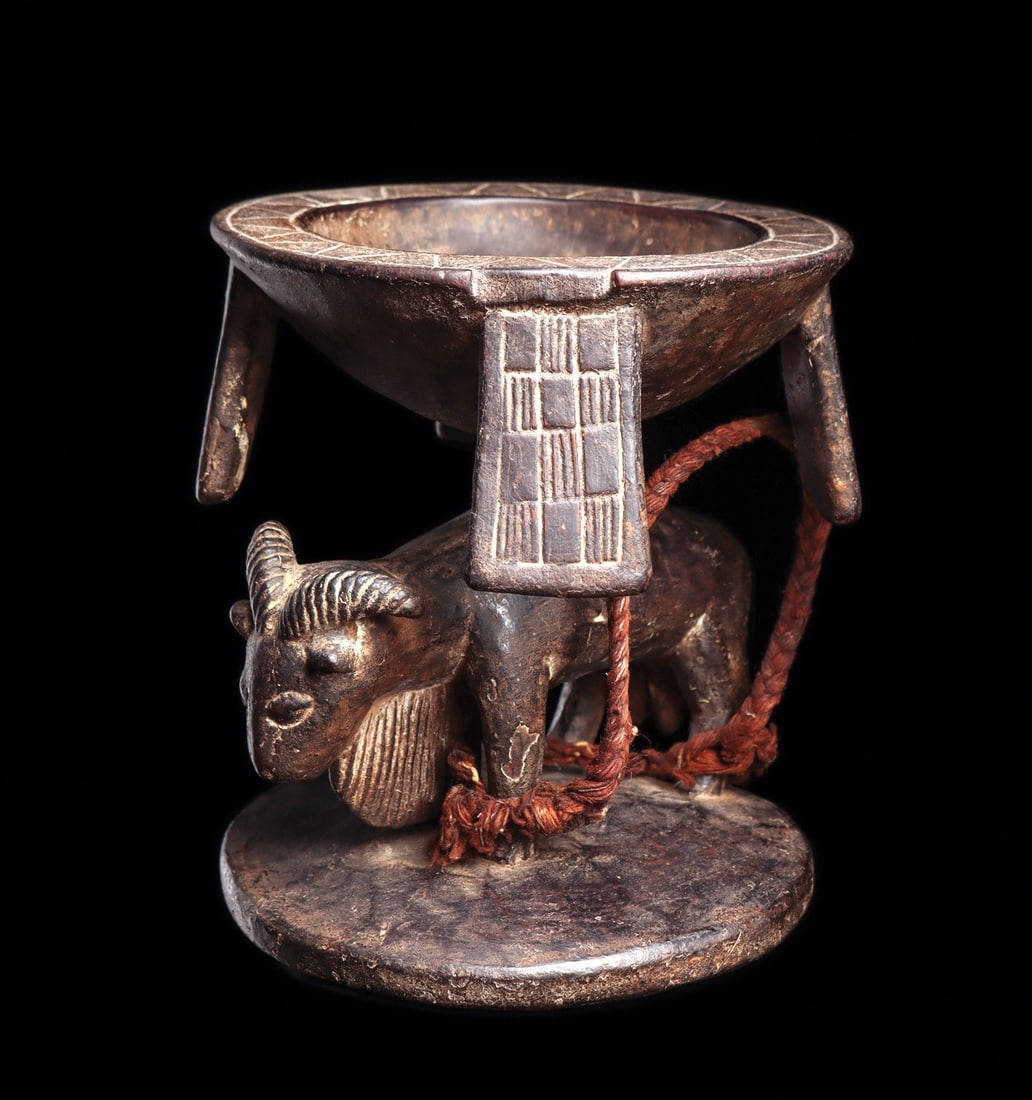 Yoruba Divination Bowl: Nigeria. Ritual bowl with an antelope with curved and grooved horns and large dewlap hanging between front legs on integral disc base. Four panels hang at edges of flat rim bowl. Dark brown patina
