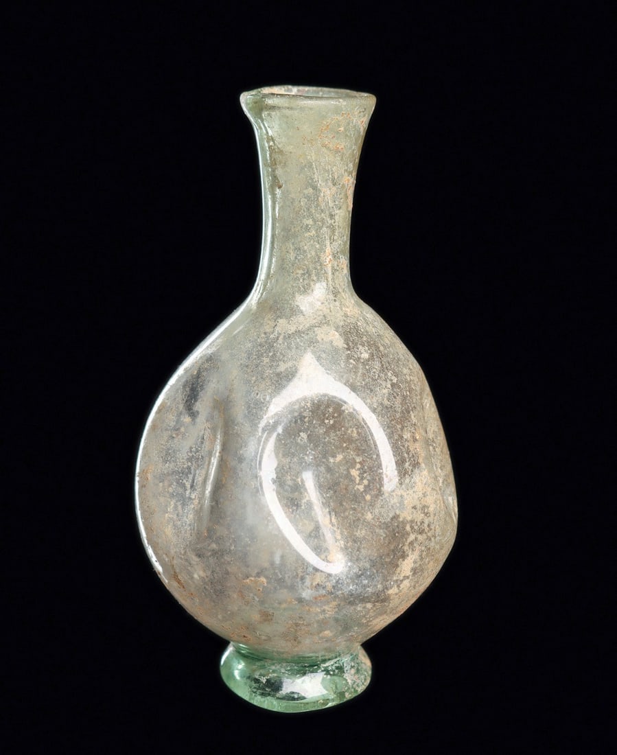Roman Vertical Buckled Glass Vessel: Eastern Mediterranean. Ca. 2nd - 3rd century CE. Light yellow-green blown glass bottle, having a tapered bulbous body decorated with five indented buckles, tapered cylindrical neck with a sheared, fir