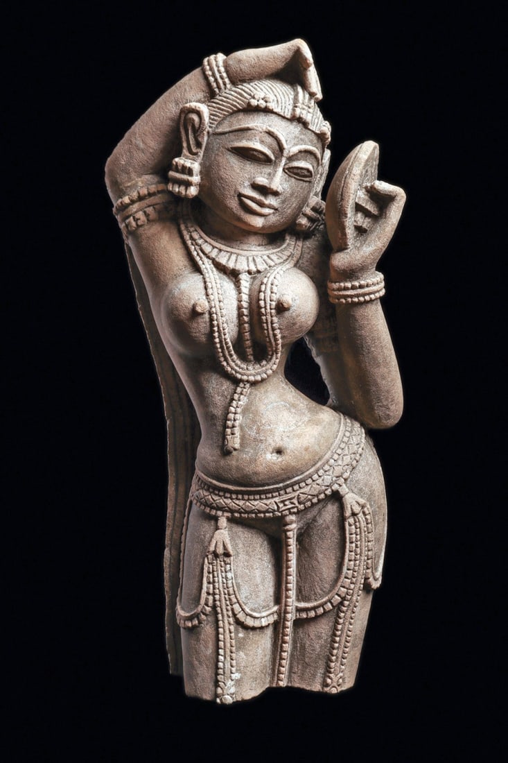 Apsara Sandstone Decorative Figure: India. Ca. 20th century. Modern carved sandstone female figure from a frieze, depicting an Apsara figure having prominent round bare breasts, and wearing a long beaded necklace with a multi-pendant co