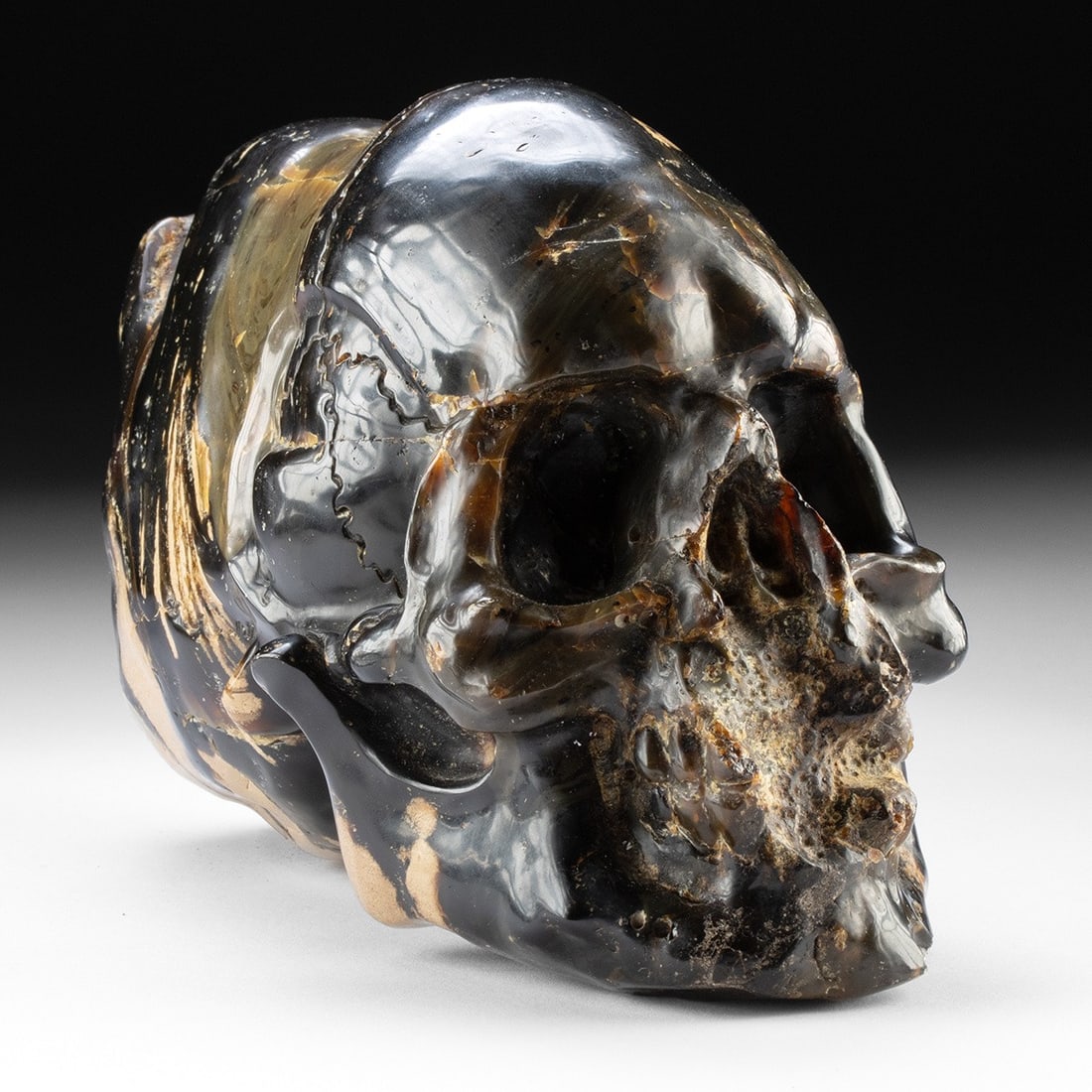 Indonesian Amber Carved Human Skull: A human skull emerges from a raw amber nodule as though the carver thought it impolite to disturb the back half of a material this beautiful, leaving the cranium's rear as nature formed it while coaxi