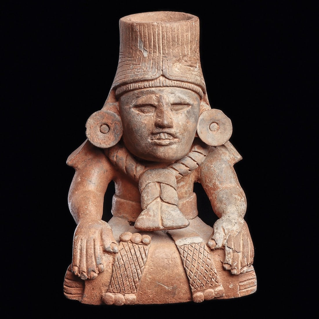 Zapotec Seated Figural Urn: Monte Alban, ca. 500 - 750 CE. Grayware pottery vessel depicting a seated god with hands held to his knees, wearing a striated headdress with side lappets, large ear discs and rope style necklace with