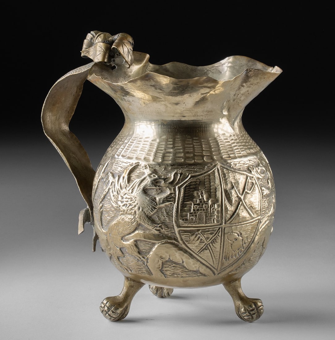 Large Peruvian Brass Pitcher w/ Heraldic Motifs: South America, Peru or Bolivia., ca. 1900 - 1966 CE. A Colonial Revival style nickel brass pitcher with a rounded body raised on three paw feet and a flared, undulating rim. The surface is worked in r