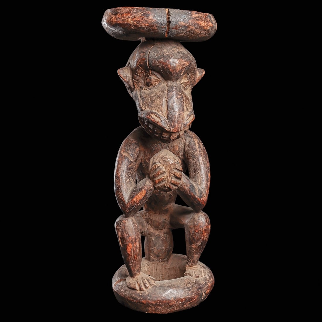 Grassfields Monkey Stool: Cameroon, . Caryatid figure of a seated monkey figure holding an ovoid form, possibly a coconut. The animated head is carved with a projecting snout, wide mouth with boldly defined teeth, bulging eyes