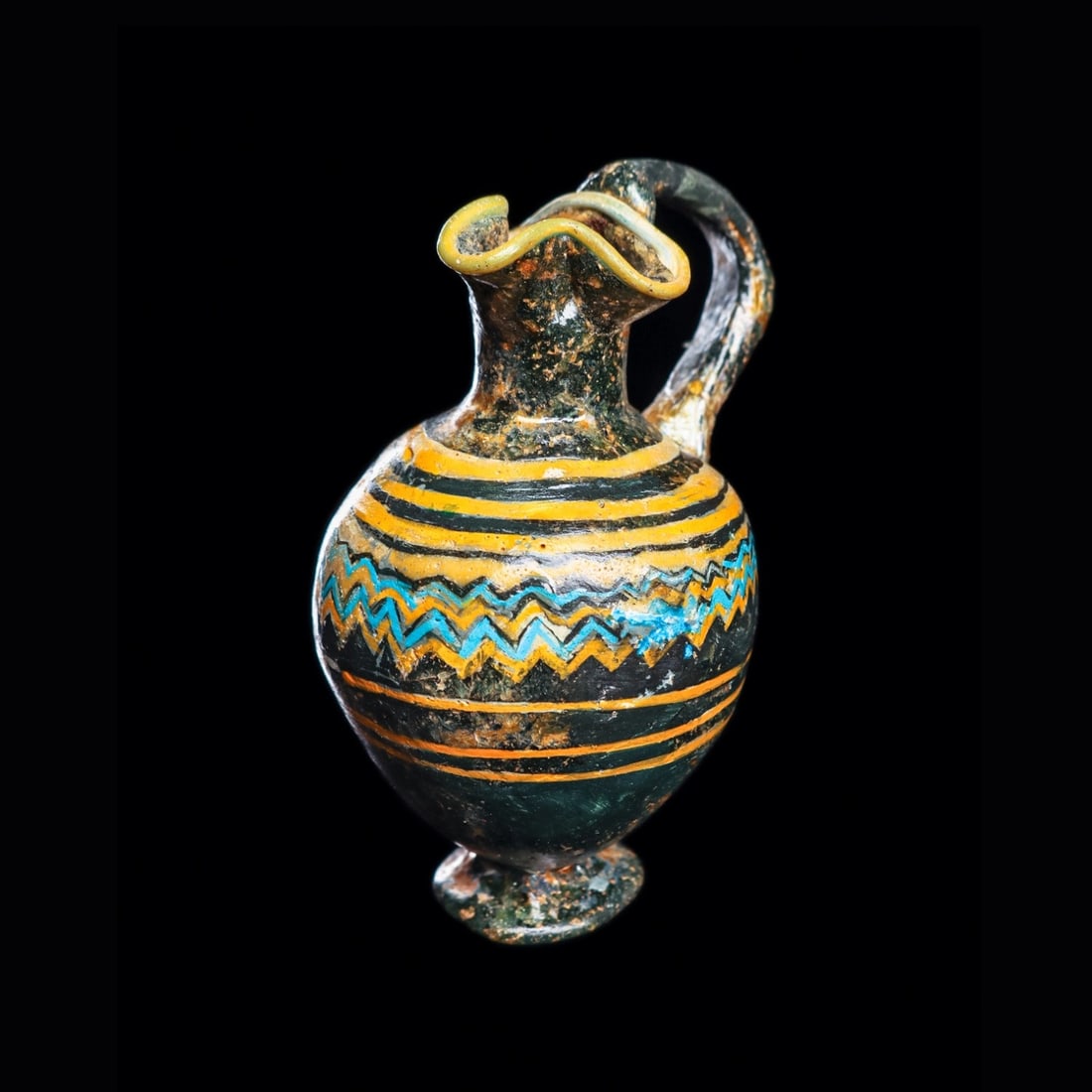 Phoenician Core-Formed Glass Oinochoe: Eastern Mediterranean to Middle East, ca. 5th - 4th century BCE. Core-formed paste glass oinochoe having a trefoil lip, applied ear shape handle and small disc foot. Decorated with light blue and