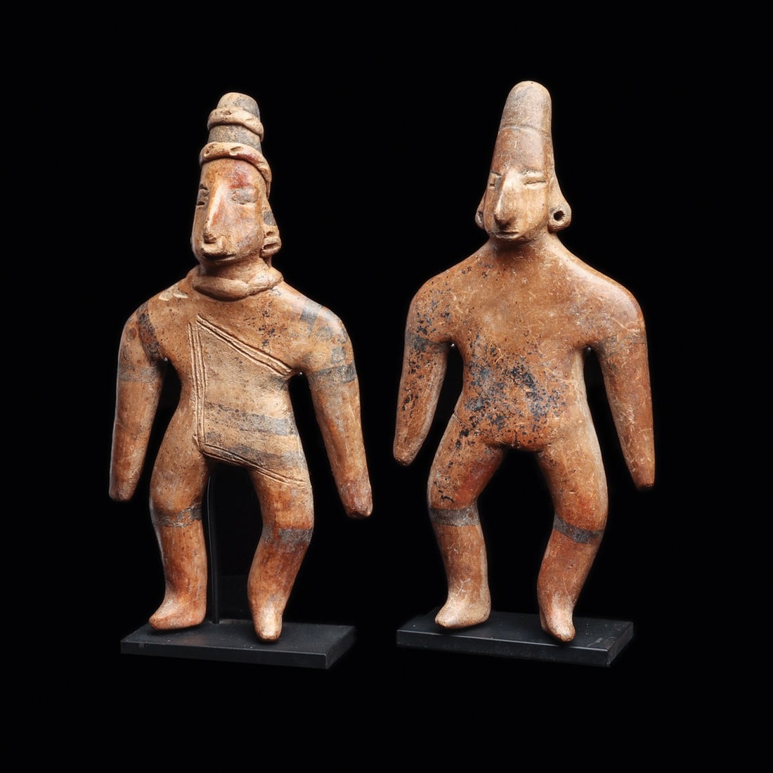 Colima Standing Gingerbread Figures (2): Colima, ca. 100 BCE – 250 CE. Two solid tan color pottery standing gingerbread figures, both having elongated pointed heads. One wearing a rope type headband, nose ring, ear ornaments and neckla