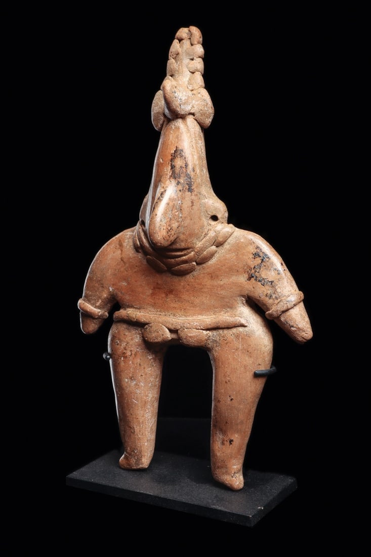 Colima Large Standing Gingerbread Figure: Colima, ca. 100 BCE – 250 CE. Large solid salmon-tan color pottery standing figure having a elongated pointed head, wearing a horned headdress, double strand beaded necklace, bracelets and belt.